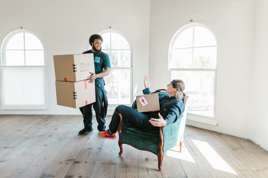 A young woman sitting on a wooden floor surrounded by numerous cardboard moving boxes of various sizes, some sealed with red tape and others labeled with 'fragile' stickers. She is inside a room with
