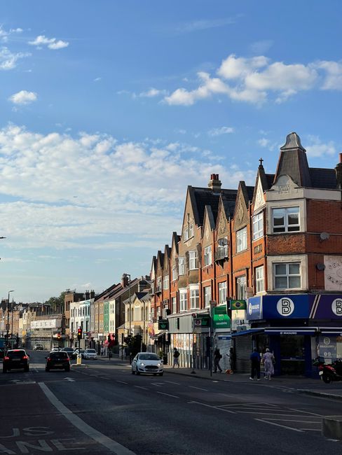 A street view of a residential area in Westfield Shepherds Bush with multi-storey brick houses featuring bay windows and pitched roofs. Several pedestrians are walking on the pavement and crossing the