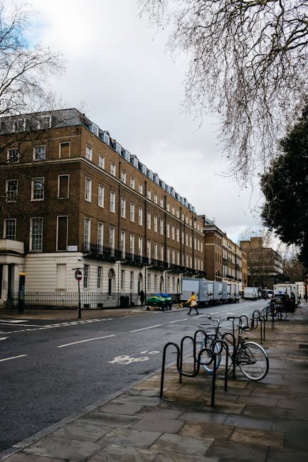 A street view of a multi-story brick residential building with white window frames and balconies, situated on Talbot Road near Brook Green. The pavement in front is lined with several bicycles secured