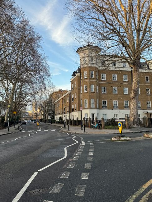 A street view of a multi-story brick residential building with white window frames and balconies, situated on Talbot Road near Brook Green. The pavement in front is lined with several bicycles secured